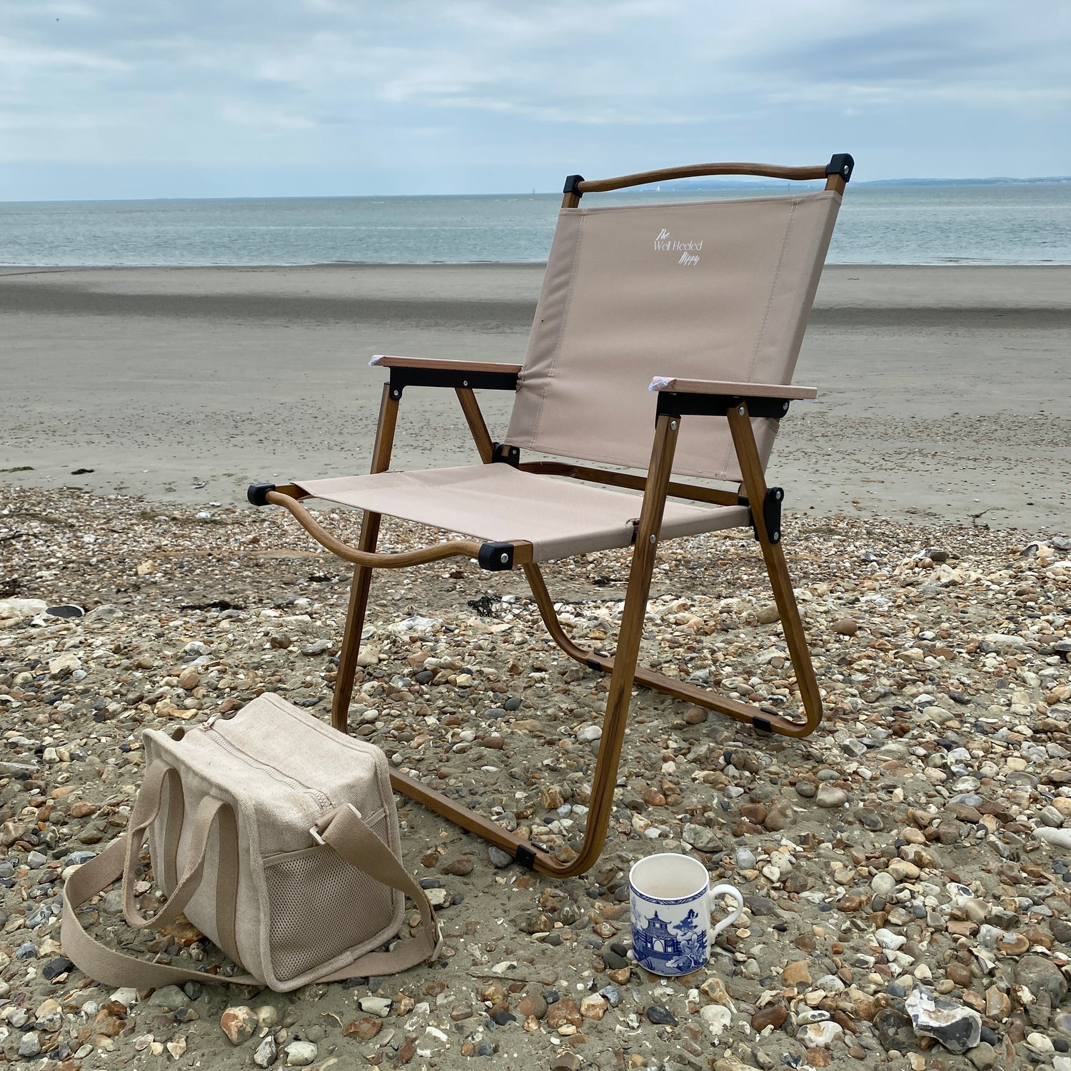 A lightweight, foldable, stylish camping chair by The Well Heeled Hippy, pictured here as a picnic chair on a pebbly beach, in beige