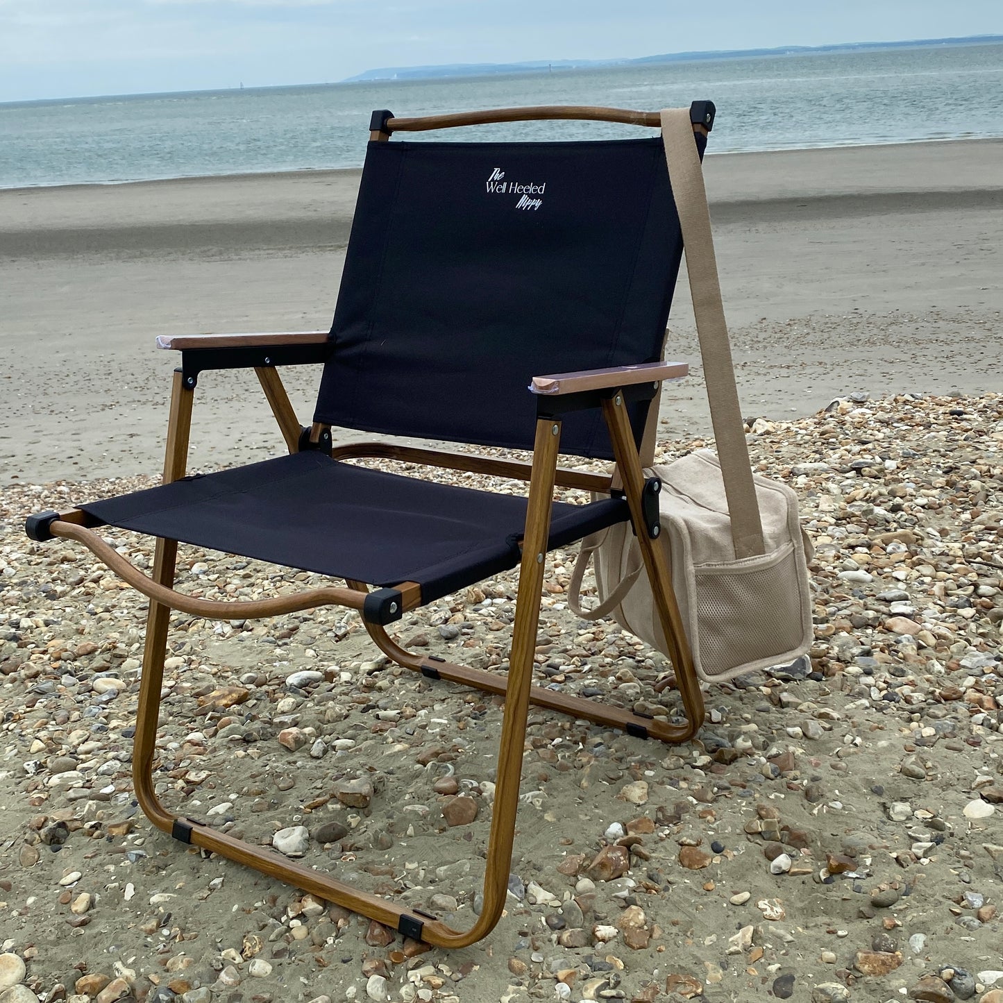 A lightweight, foldable, stylish camping chair by The Well Heeled Hippy, pictured here as a picnic chair on a pebbly beach, in black