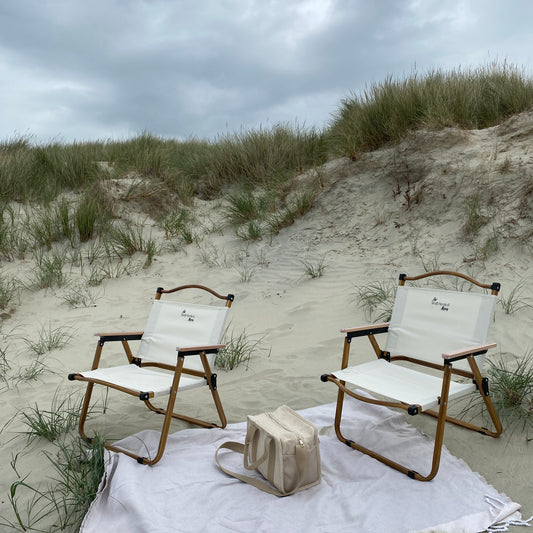 A pair of lightweight, foldable, stylish camping chairs by The Well Heeled Hippy, pictured here as a picnic chair on a sandy beach, in beige